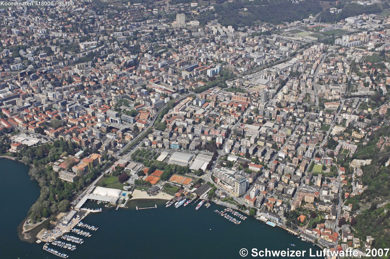 Lugano, Lido (im Vordergrund) mit Hafenanlage und Halle. Centro Esposizioni Padiglione Conza untere Bildmitte. Fluss Cassarate durchquert das Bild von oben rechts nach unten links, d.h. von Nord nach Süd. Campus der Universität: Bildmitte rechts des Flusses. Obere rechte Bildecke: Sportanlagen Cornaredo (Position 2'717'948.91, 1'098'006.23).